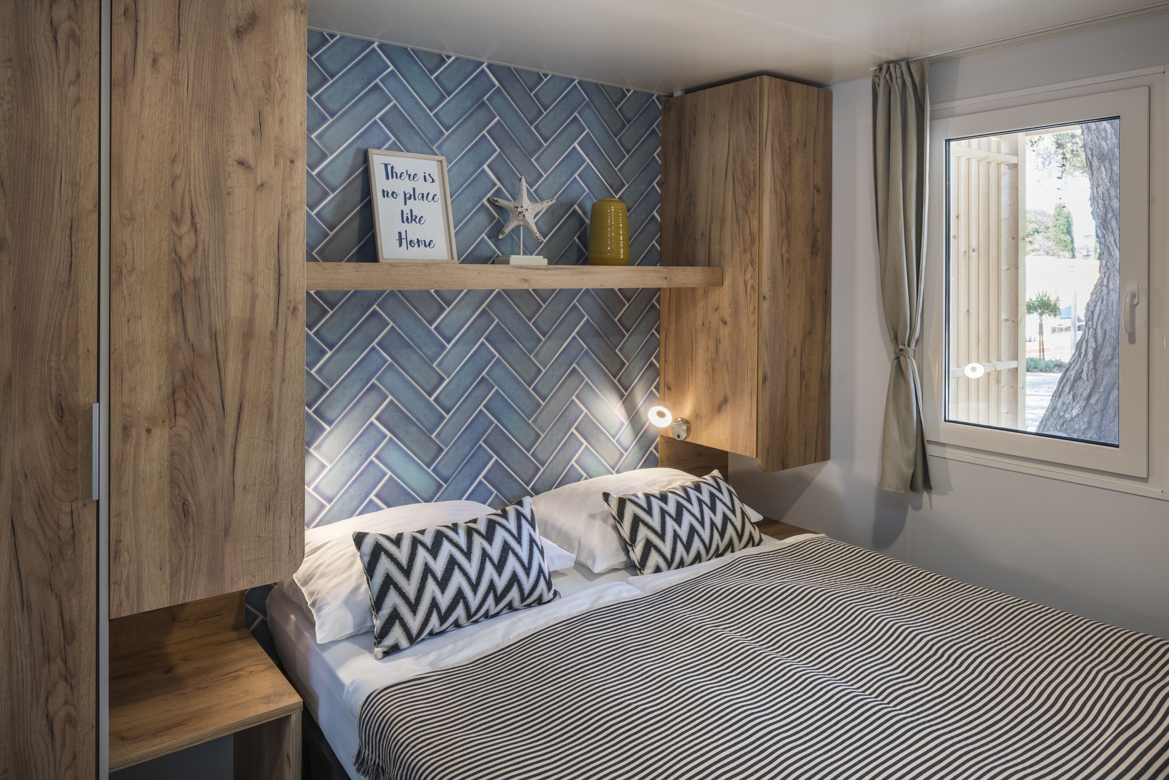 A bedroom with a herringbone-patterned wall, a bed with striped bedding, a nightstand, a shelf with decorative items, and a window with curtains.