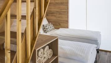 A wooden staircase leading to a bedroom with two beds, a shelf with rolled towels, and a window with blinds.