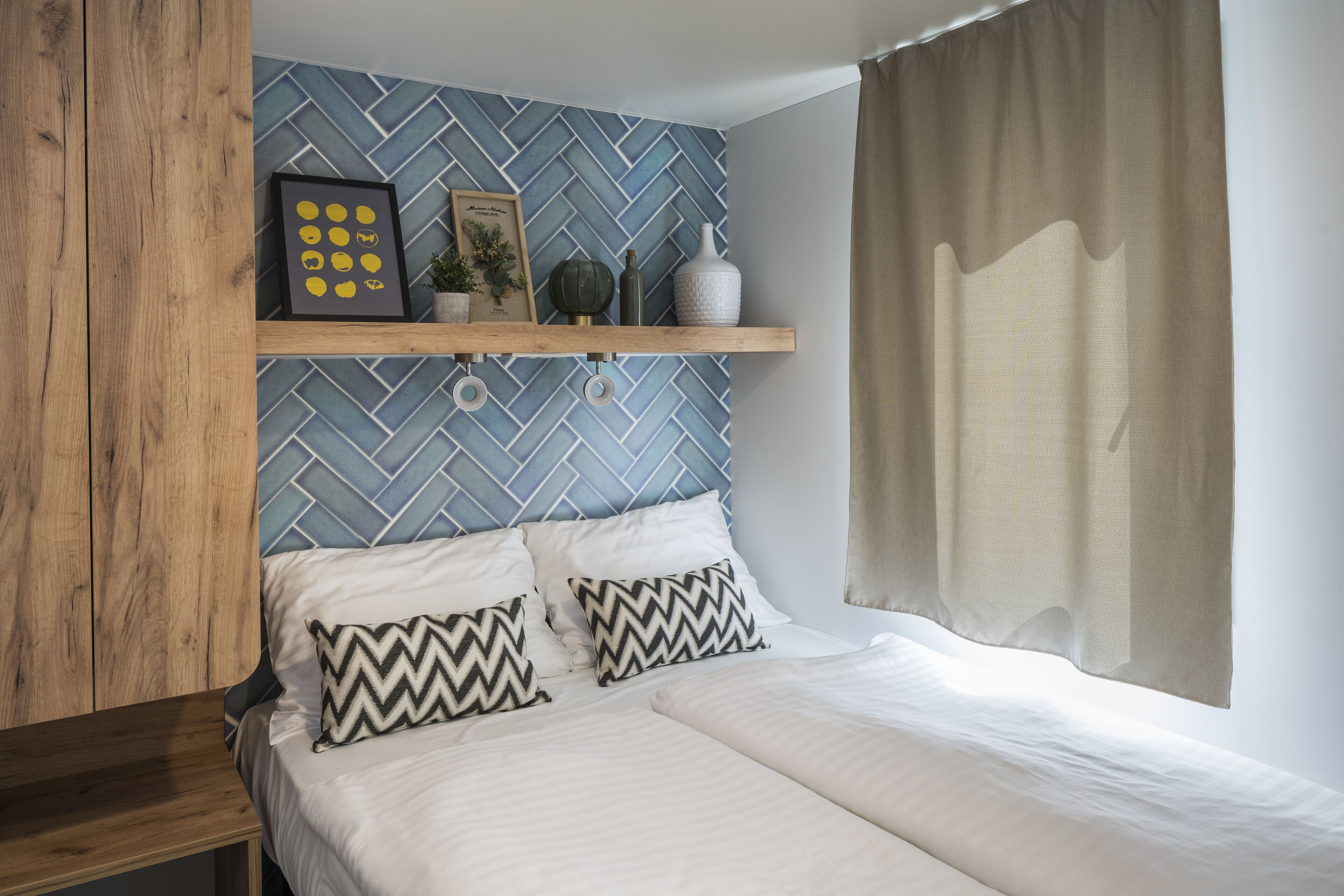 Un dormitorio con cama, dos almohadas, cabecera de madera, pared con diseño azul y blanco, repisa con objetos decorativos y ventana con cortinas.