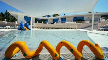 Zona de piscina con un edificio moderno, un tobogán y juguetes inflables amarillos y azules.