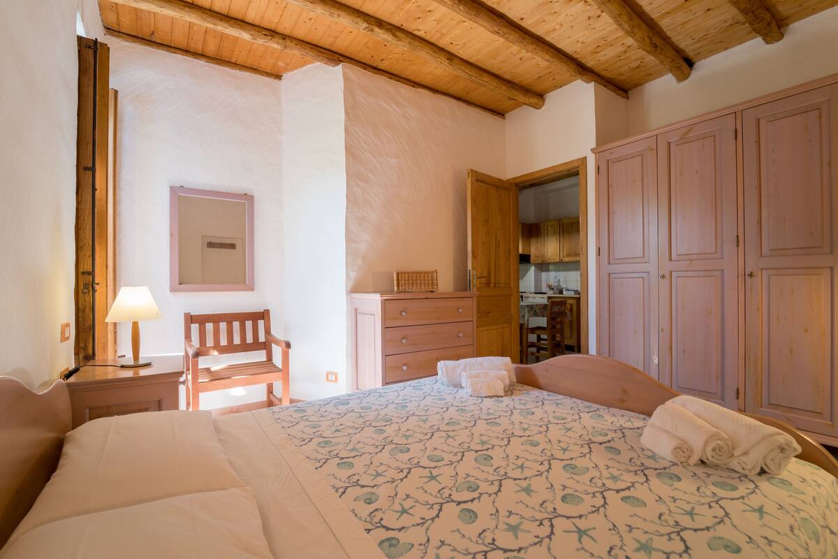 Quarto com cama, guarda-roupa de madeira, uma cadeira, escrivaninha com abajur e espelho na parede.