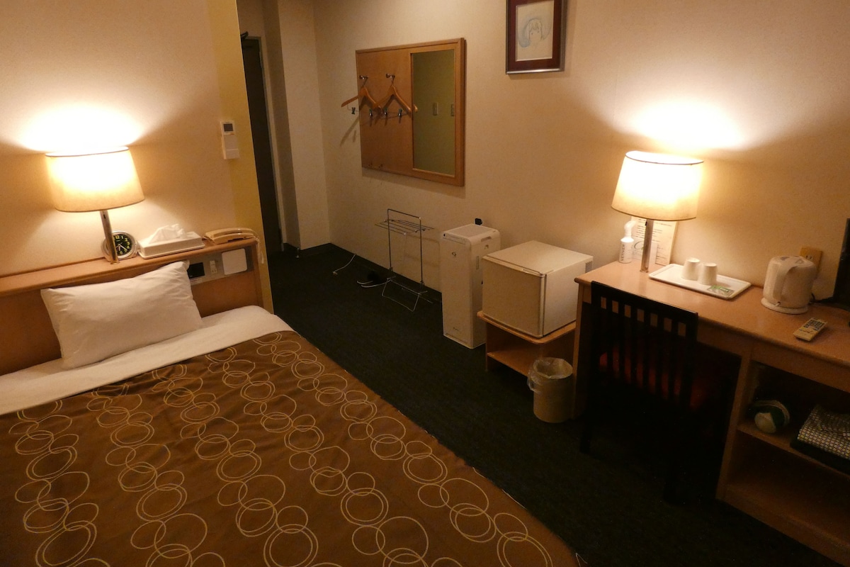 A hotel room with a bed, two bedside lamps, a desk with a chair, a small refrigerator, a waste bin, and a mirror on the wall.