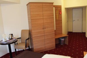 A hotel room with a wardrobe, a small table with a kettle, a chair, a bed, and a carpeted floor.