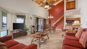 A spacious living area with a large TV, red sofas, a wooden coffee table, and a ceiling fan. - Aloha Condos, Kanaloa at Kona, Condo 3703, Oceanfront, AC (Kailua Kona)