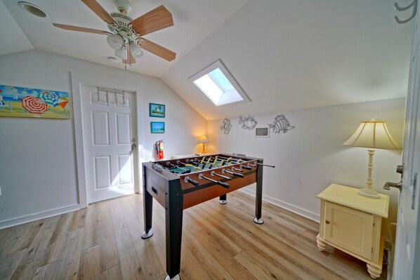A room with a foosball table, a ceiling fan, a skylight, and a small cabinet with a lamp.