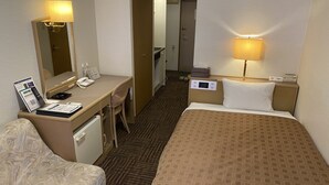 A hotel room with a bed, a desk with a telephone and a laptop, a chair, a bedside table with a lamp, and a mirror.