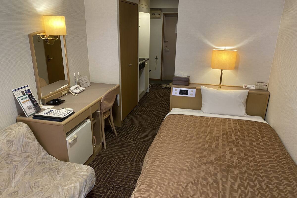 A hotel room with a bed, a desk with a telephone and a laptop, a chair, a bedside table with a lamp, and a mirror.