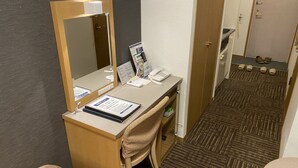 A hotel room with a desk, chair, and a large mirror. There is a telephone, a brochure, and a small trash bin. The room has a carpeted floor and a door leading to another room.