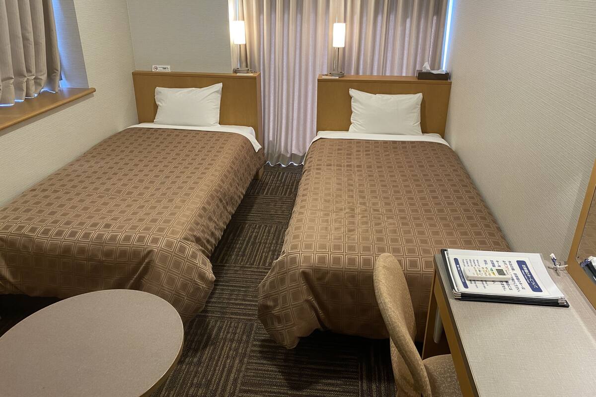 A hotel room with two single beds, a small round table, a chair, and a desk with a telephone and a menu.