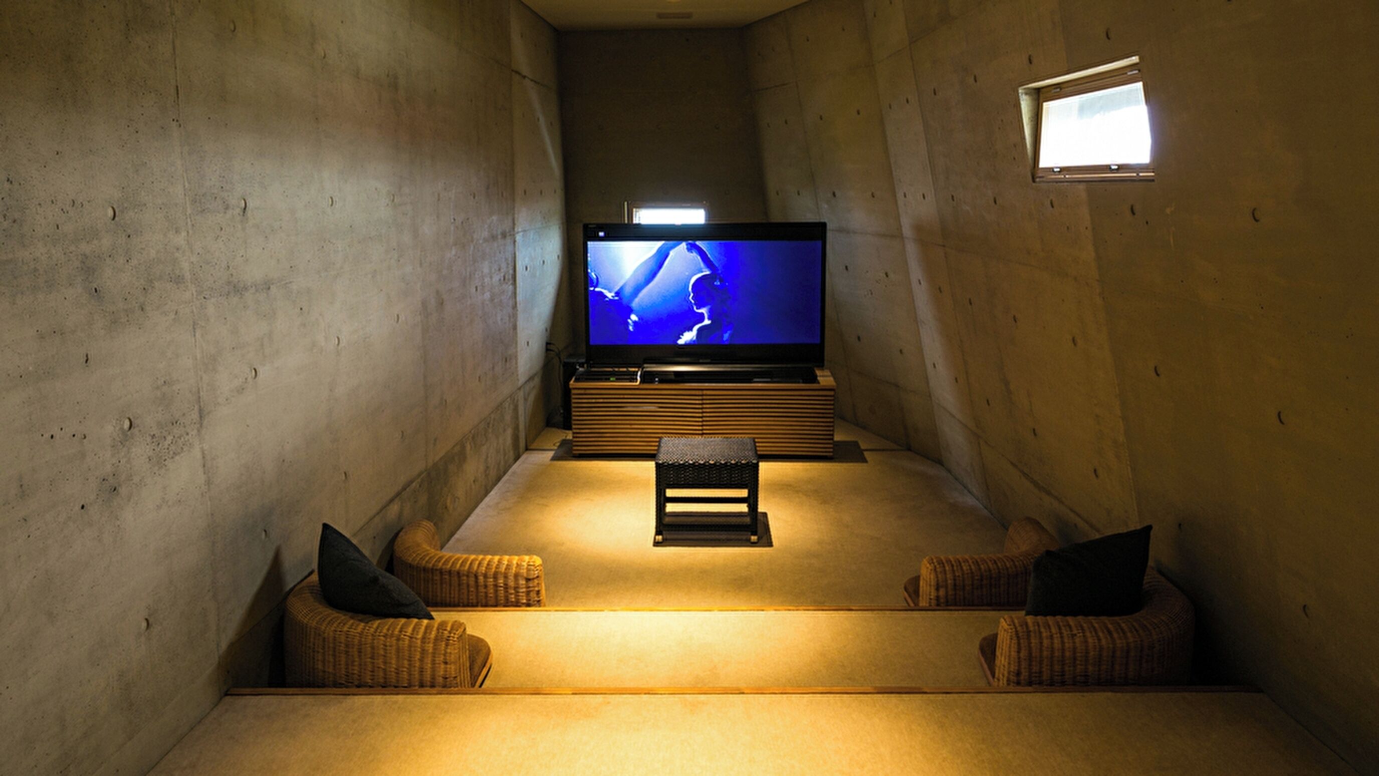 A cozy cinema room with a large flat-screen TV, a wooden bench, and two wicker chairs.