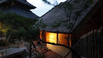 A traditional Japanese building with a thatched roof and wooden structure, surrounded by a garden with rocks and sparse vegetation.