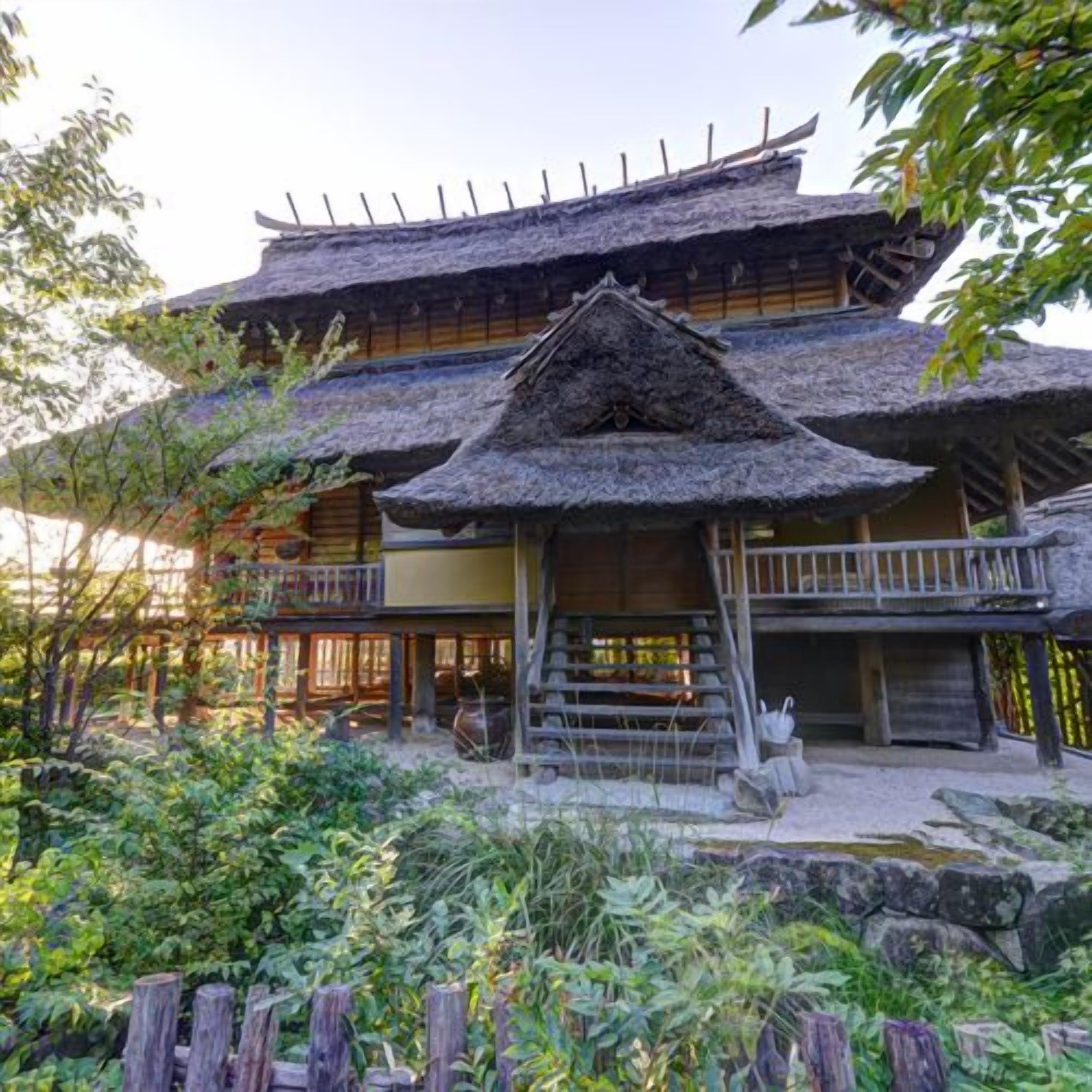 A traditional Japanese-style building with a thatched roof, wooden balconies, and a garden area with rocks and plants.