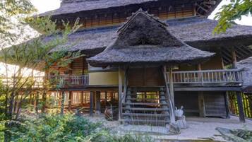 A traditional Japanese-style building with a thatched roof, wooden balconies, and a garden area with rocks and plants.