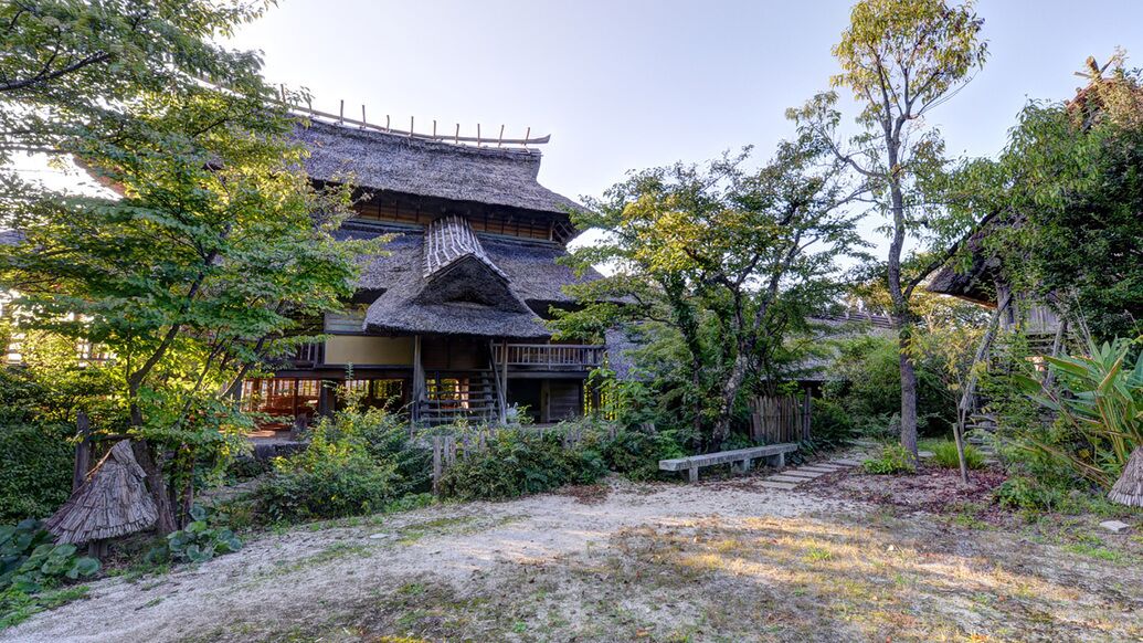 Un edificio japonés tradicional con techo de paja, rodeado de exuberante vegetación y una terraza de madera.