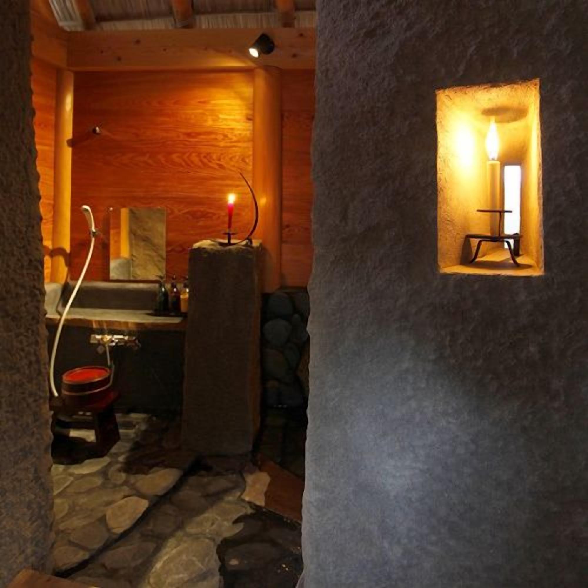A rustic bathroom with a stone floor, wooden walls, a candle in a niche, and a small sink.