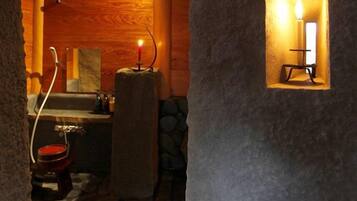 A rustic bathroom with a stone floor, wooden walls, a candle in a niche, and a small sink.
