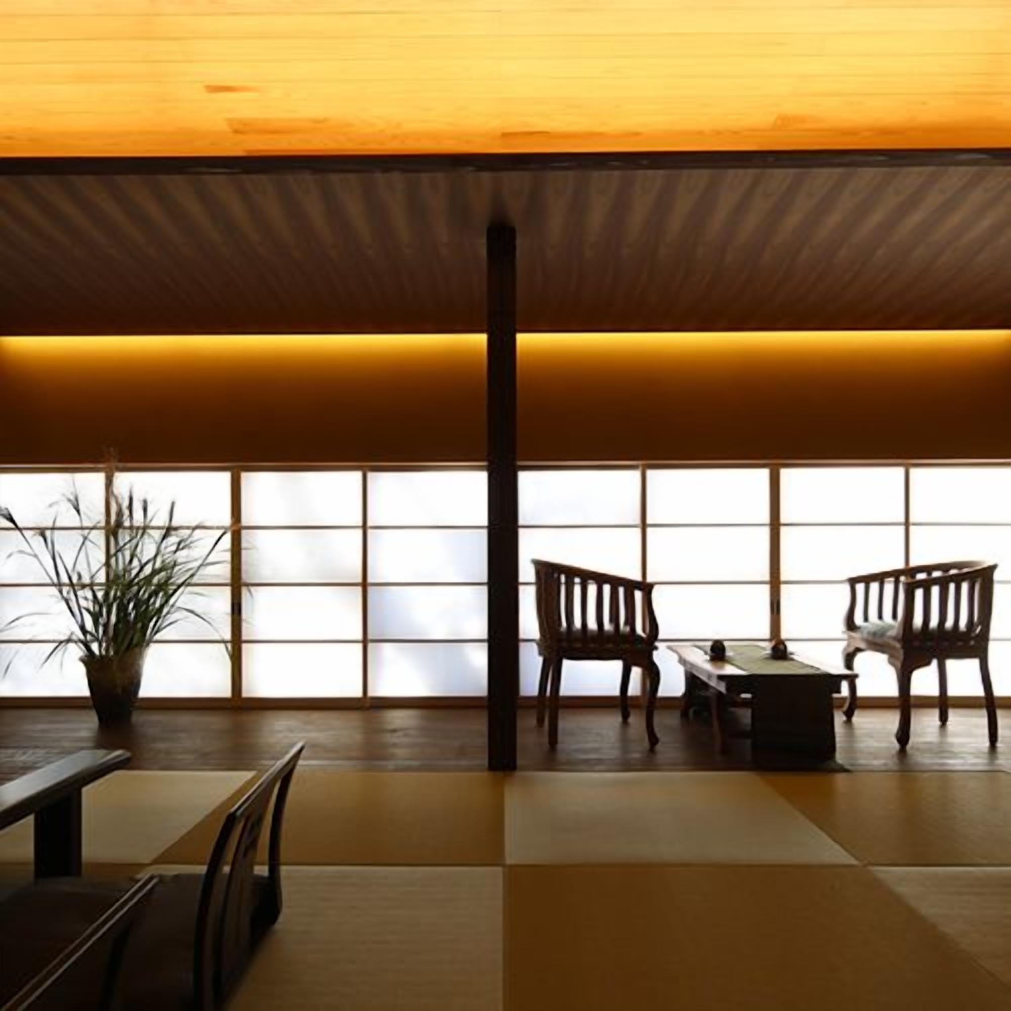 A traditional Japanese room with tatami flooring, a low table, two wooden chairs, and a potted plant.