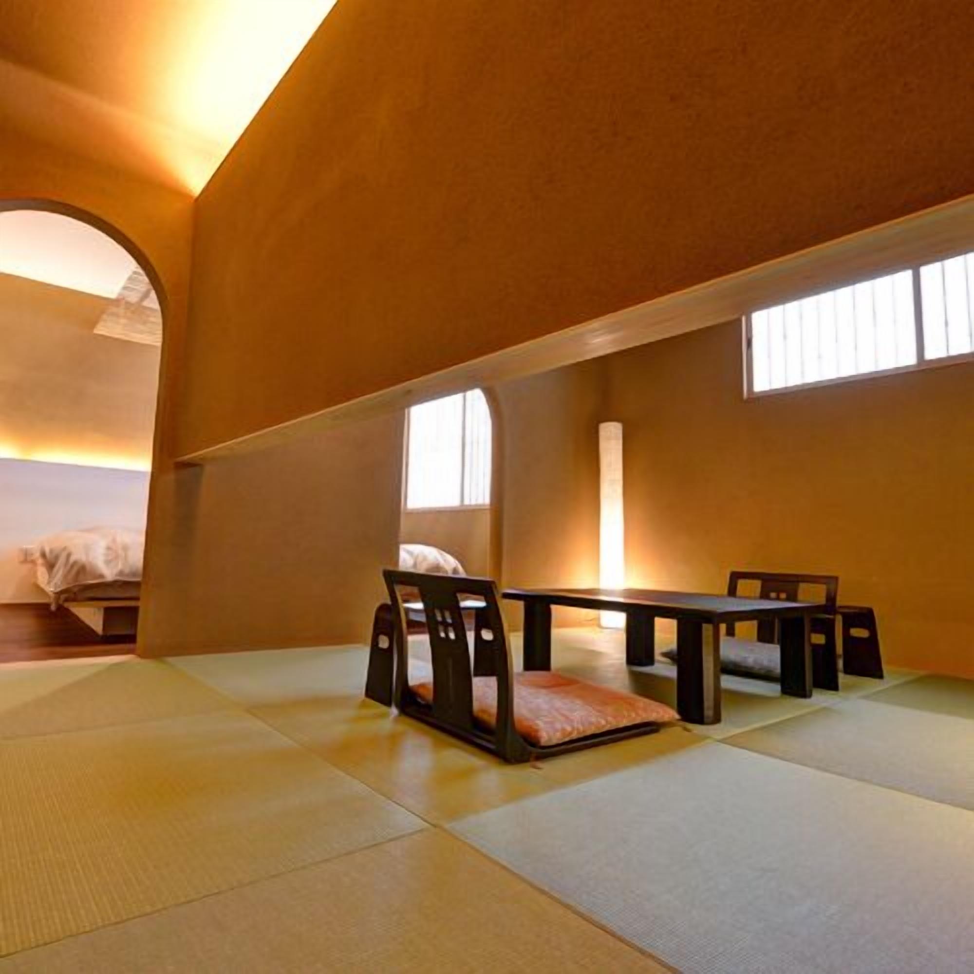 A tatami mat room with a low table and chairs, a bed in the background, and a window providing natural light.