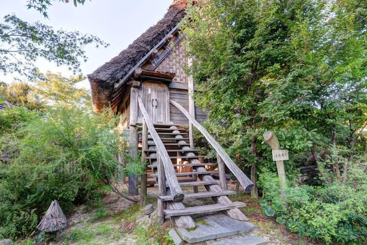 A traditional wooden staircase leading to a thatched-roof building surrounded by dense greenery.