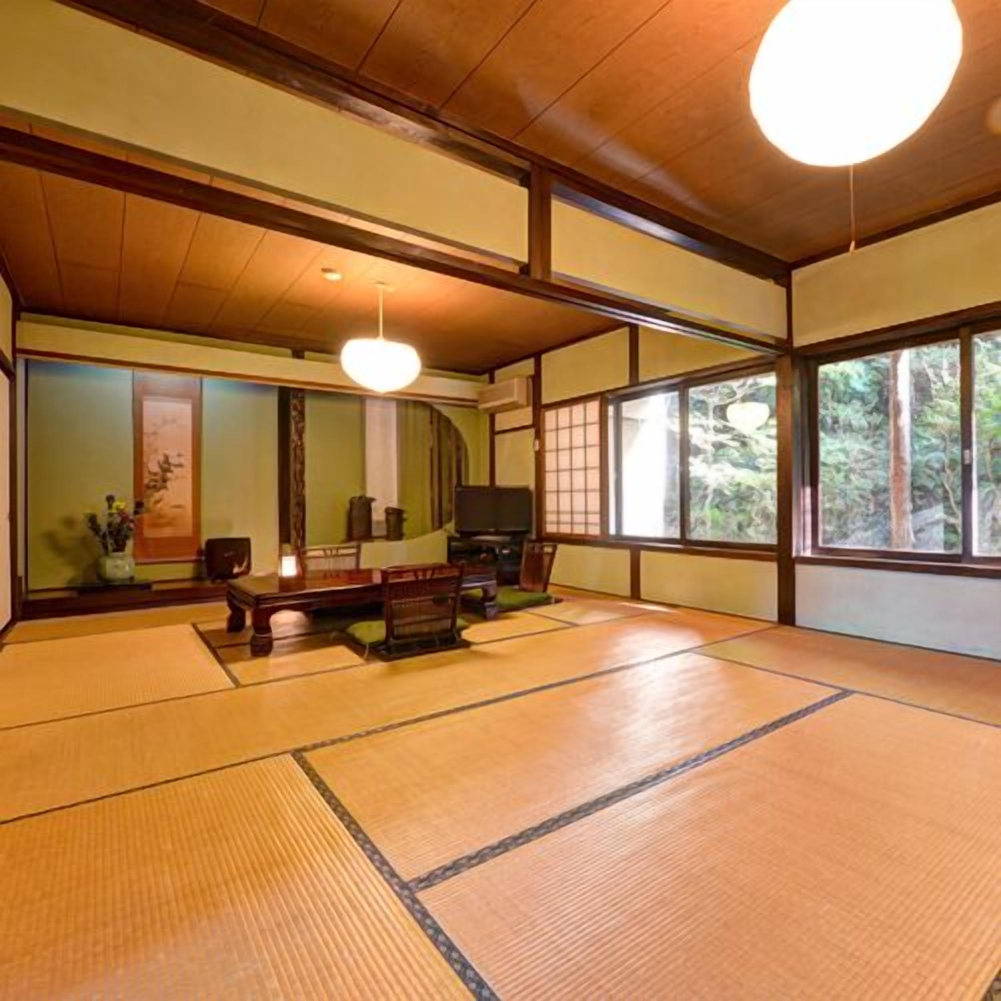 A tatami mat room with a low table, chairs, a hanging light, and a view through large windows.