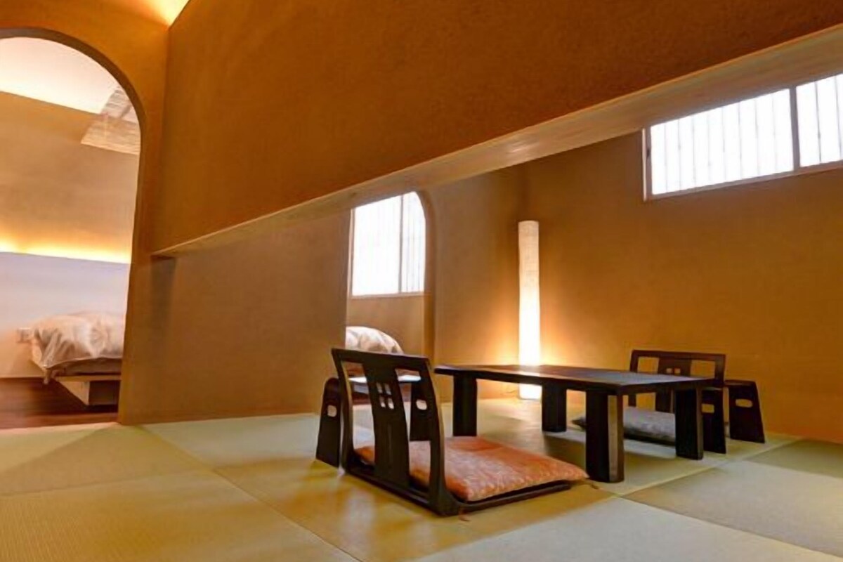 Una habitación con esteras de tatami, una mesa baja y sillas, una cama al fondo y una ventana que deja entrar luz natural.