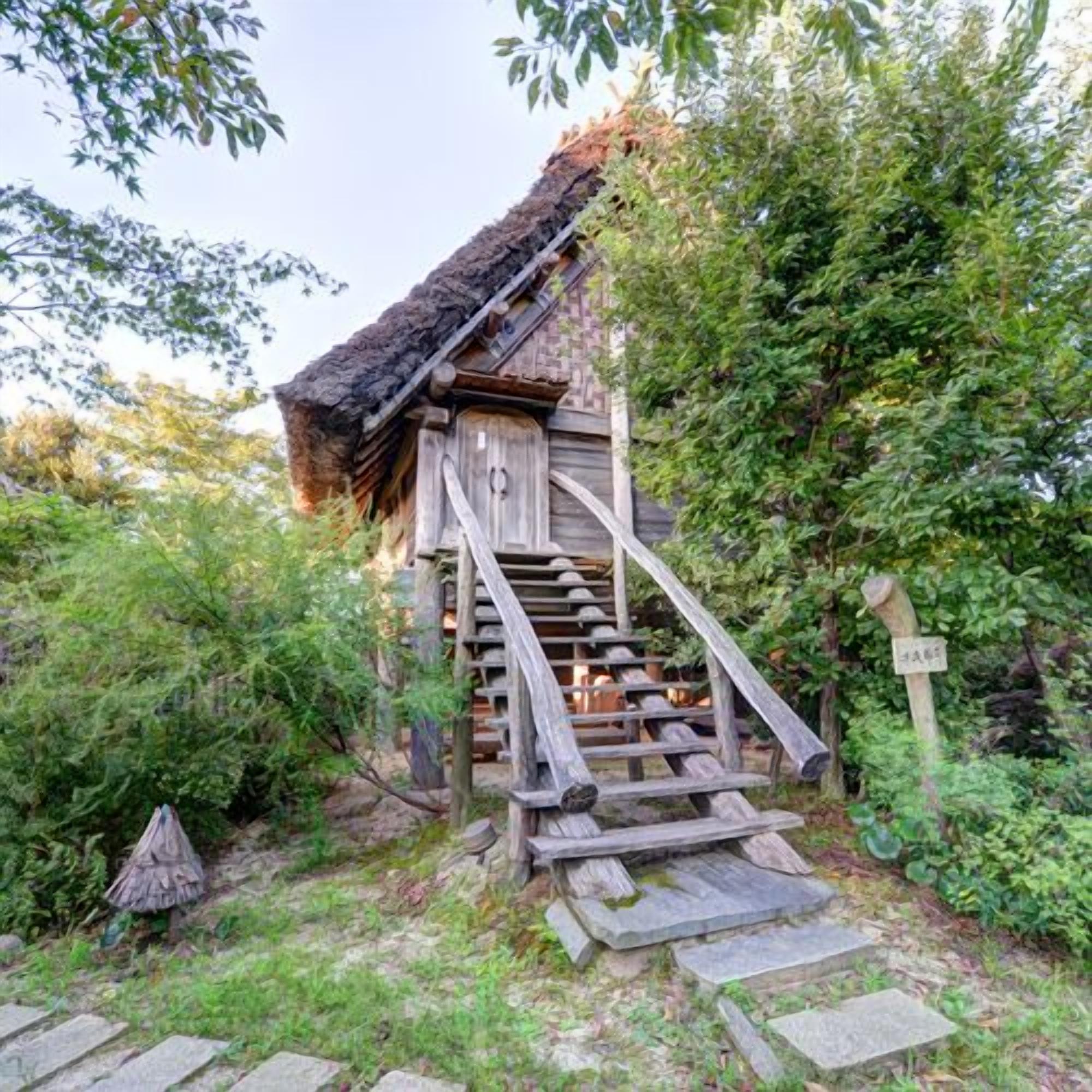 A wooden staircase leading to a rustic building with a thatched roof, surrounded by greenery.
