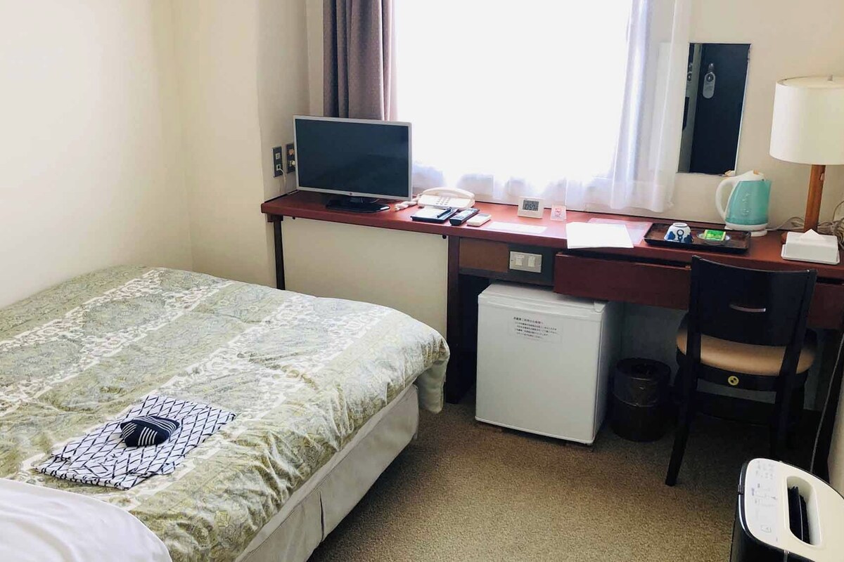 Une chambre d’hôtel comprenant un lit, un bureau avec un ordinateur, un petit réfrigérateur, une chaise et une lampe.