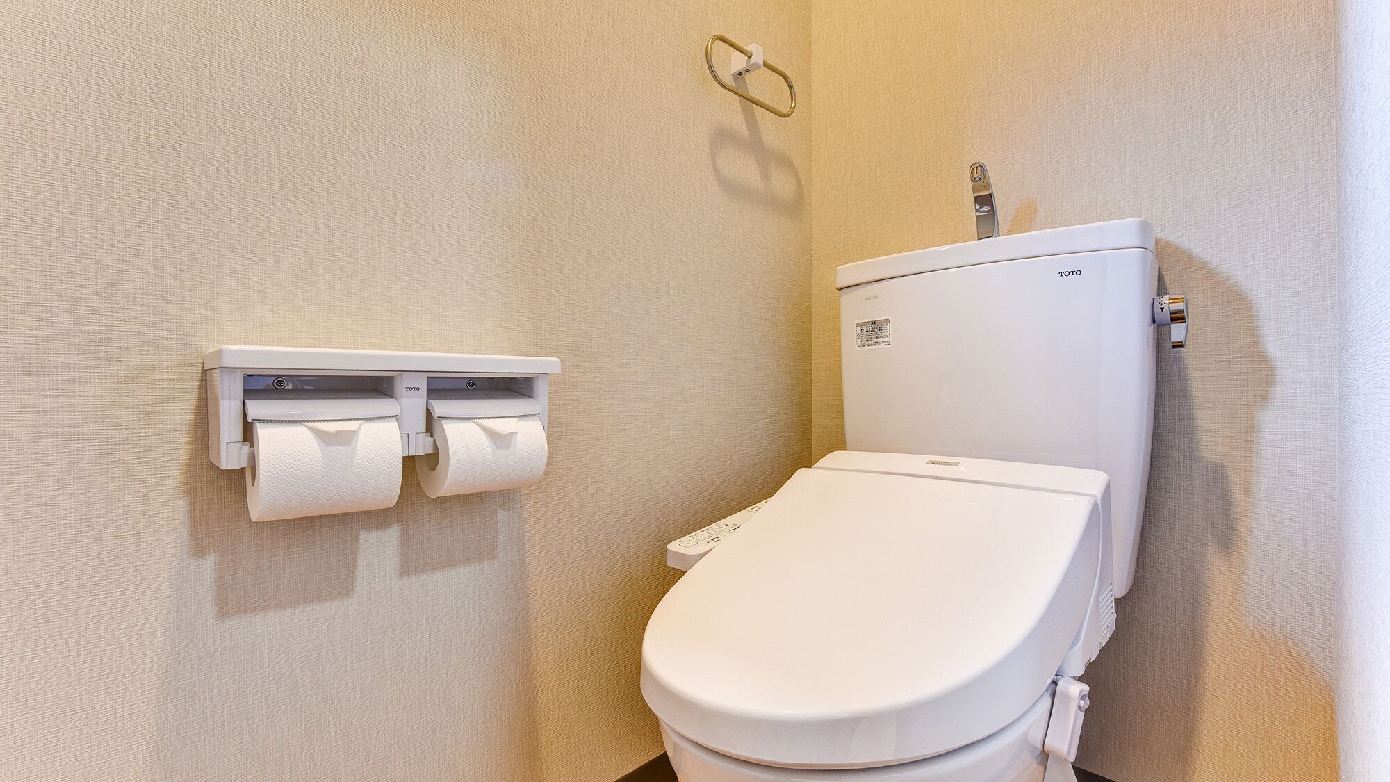Des toilettes avec un abattant et un couvercle blancs, un porte-rouleau de papier toilette fixé au mur, contenant deux rouleaux, et un distributeur de papier toilette visible.
