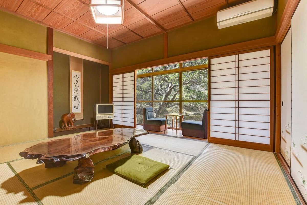 Une pièce avec un sol recouvert de tatamis, une table en bois, un coussin vert, une télévision et une porte coulissante donnant sur un paysage arboré.