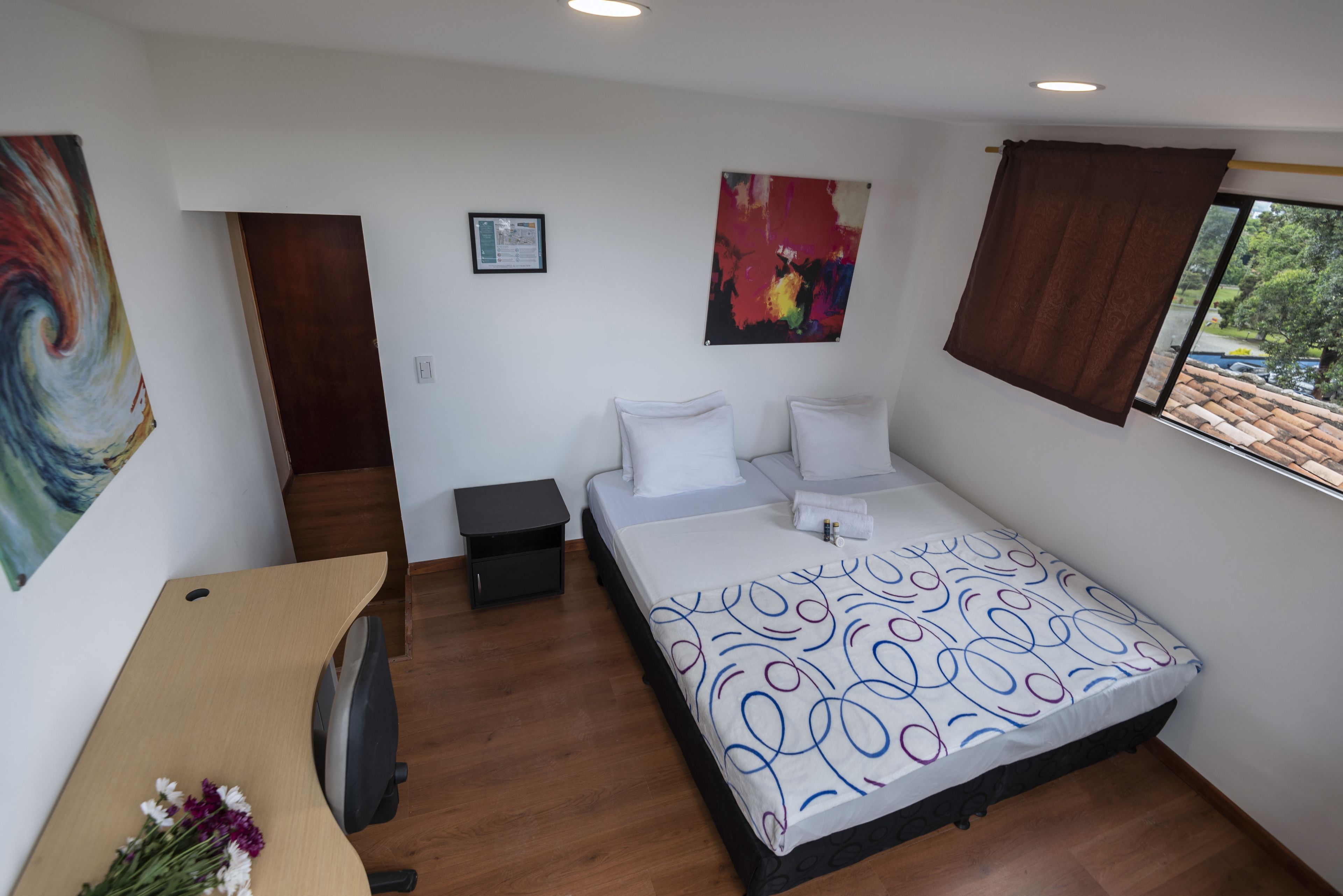 A bedroom with a bed, a desk, a chair, a small table, and a window with curtains.