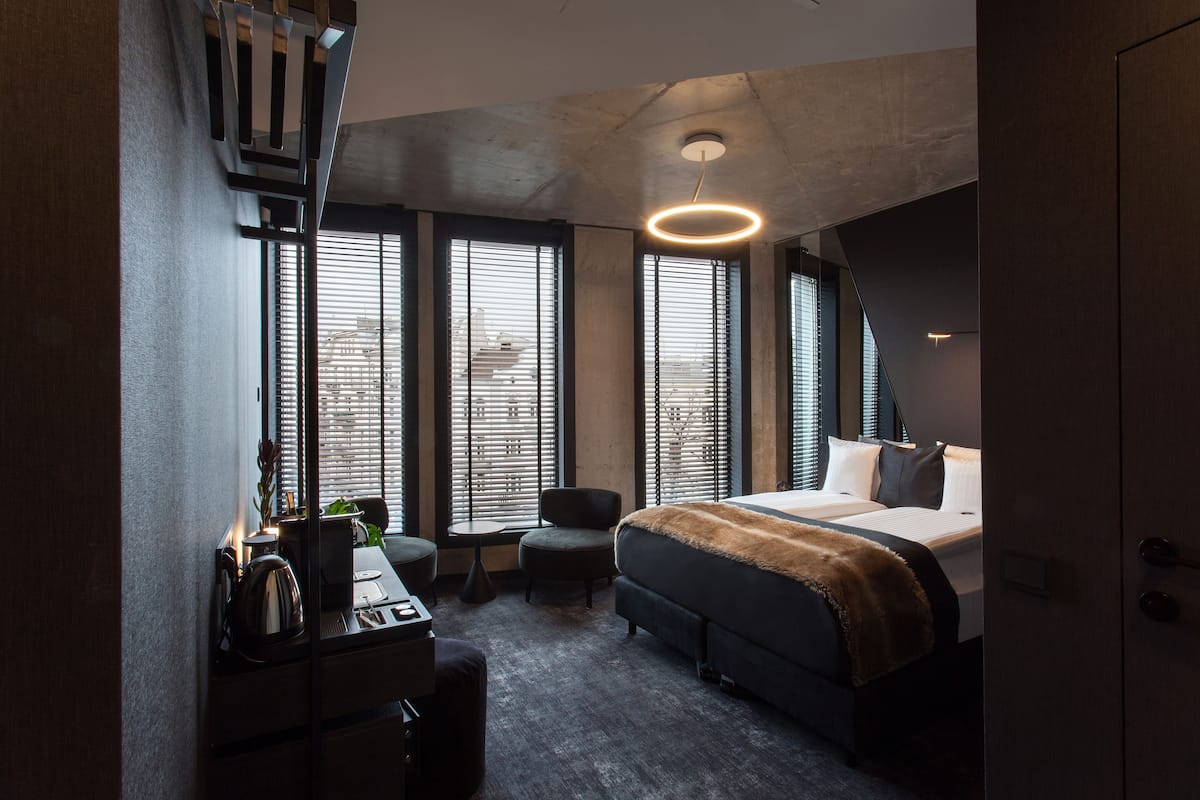 A modern hotel room with a large bed, a desk, and a chair. There are multiple windows with blinds, and a unique light fixture above the bed.