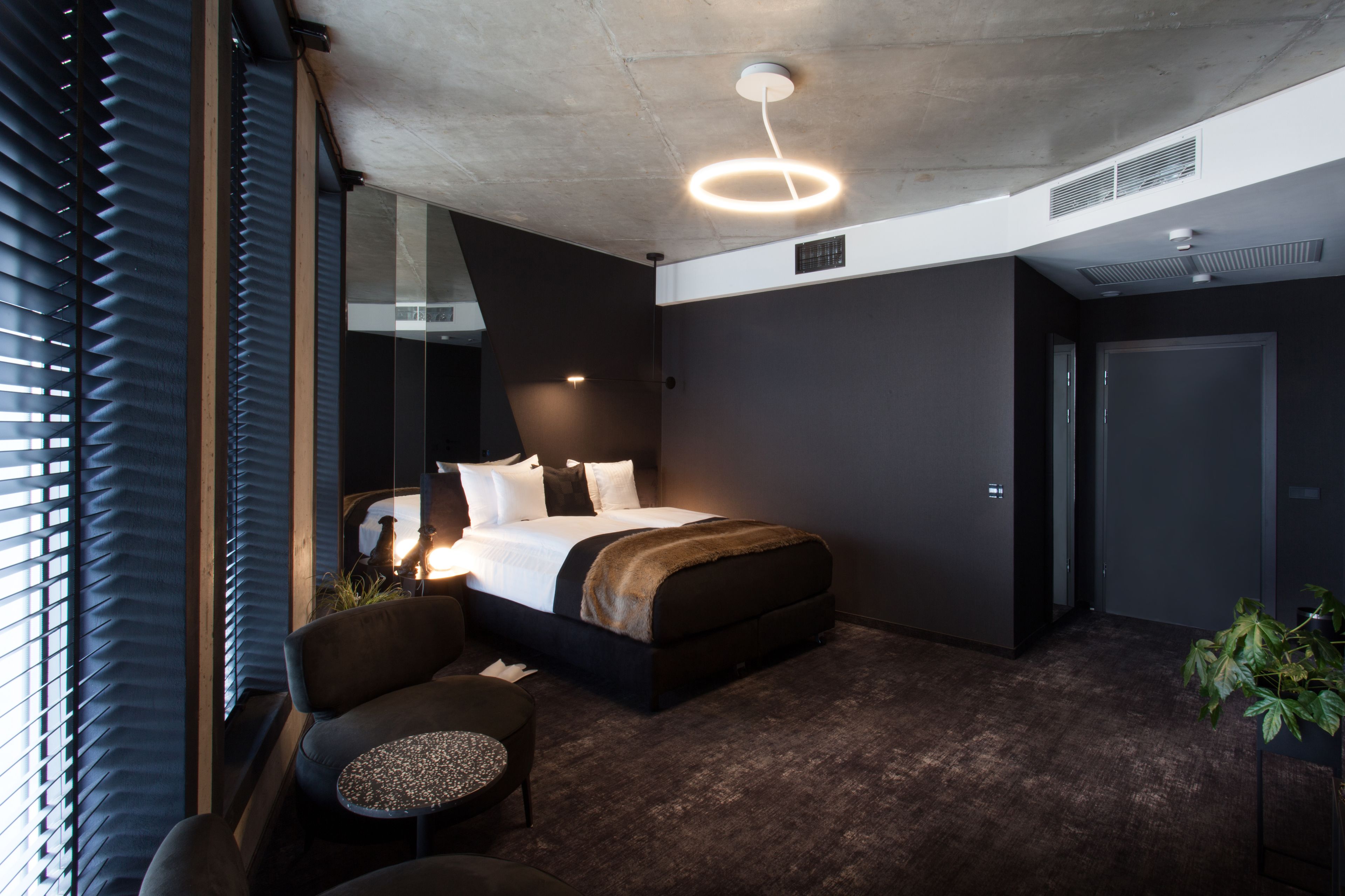 A modern hotel room with a large bed, a seating area with a small table, and a large window with blinds.