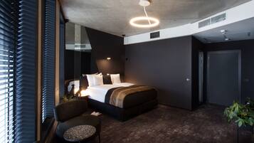 Una habitación de hotel moderna con una cama grande, una zona de estar con una mesita y un ventanal con persianas.