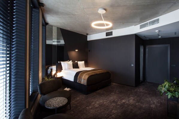 A modern hotel room with a large bed, a seating area with a small table, and a large window with blinds. - The Loft Hotel - Adults Only (Kraków)