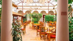 An indoor dining area with wooden tables and chairs, surrounded by columns and potted plants. - Plaza Magnolias Suites (San Cristóbal de las Casas)