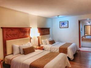 A hotel room with two beds, wooden headboards, bedside lamps, a framed picture on the wall, and a bathroom visible through an open door. - Plaza Magnolias Suites (San Cristóbal de las Casas)