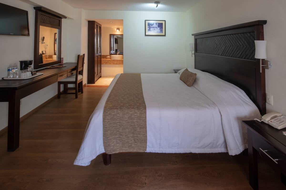 Ein Hotelzimmer mit einem großen Bett, einem Schreibtisch mit Stuhl, einem Fernseher und einem durchgegangene Tür erkennbaren Badezimmer.