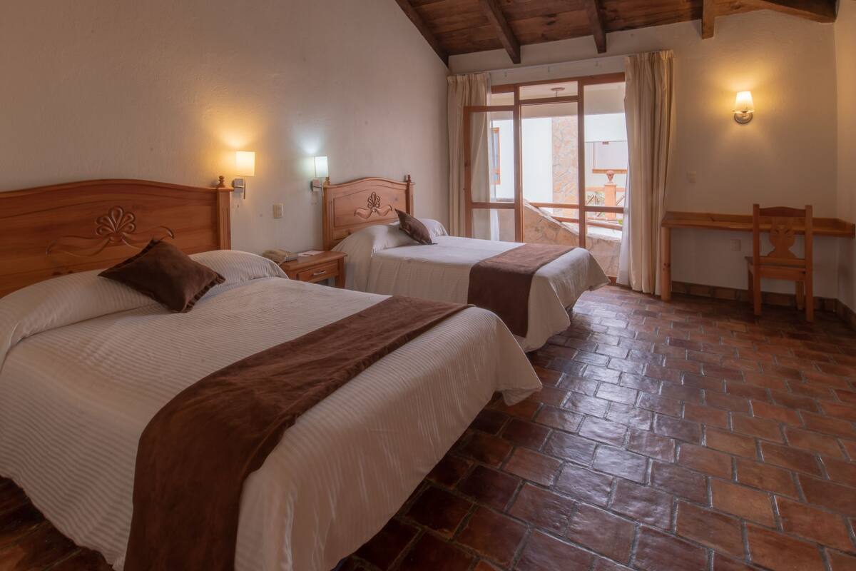Ein Zimmer mit zwei Betten, einem hölzernen Kopfteil, einem Ziegelboden und einem Balkon mit Tisch und Stuhl.