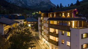 A multi-story hotel with balconies, a mountain backdrop, and a lit street. - The Peak Hochkönig (Dienten am Hochkoenig)