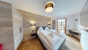 A modern bedroom with a wooden wall, a bed with white linens, a bedside table, a desk, and a large window. - The Peak Hochkönig (Dienten am Hochkoenig)