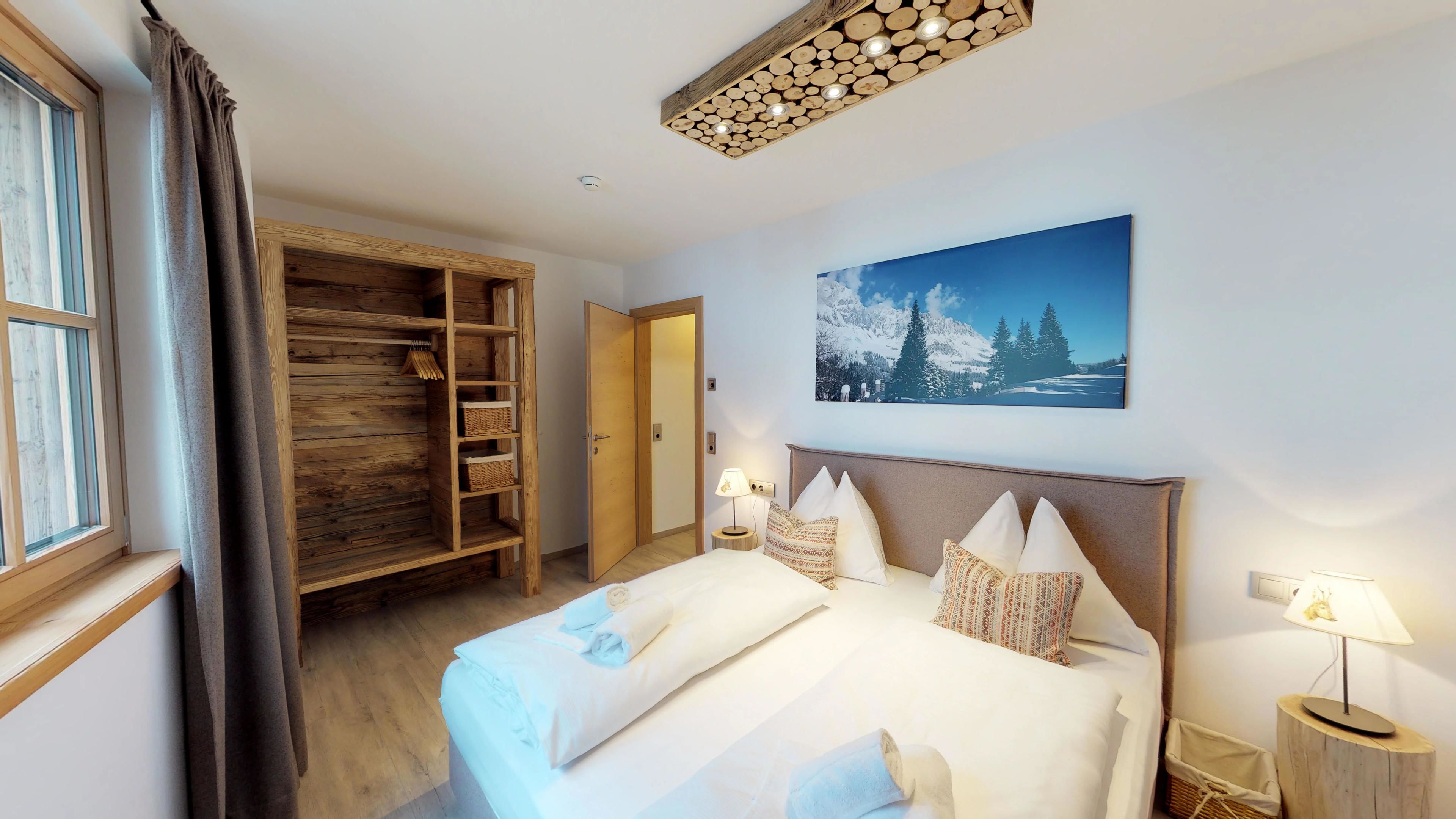 Un dormitorio con un armario de madera, una cama con almohadas, una mesita de noche, una lámpara de pared y un cuadro de una escena montañosa nevada.