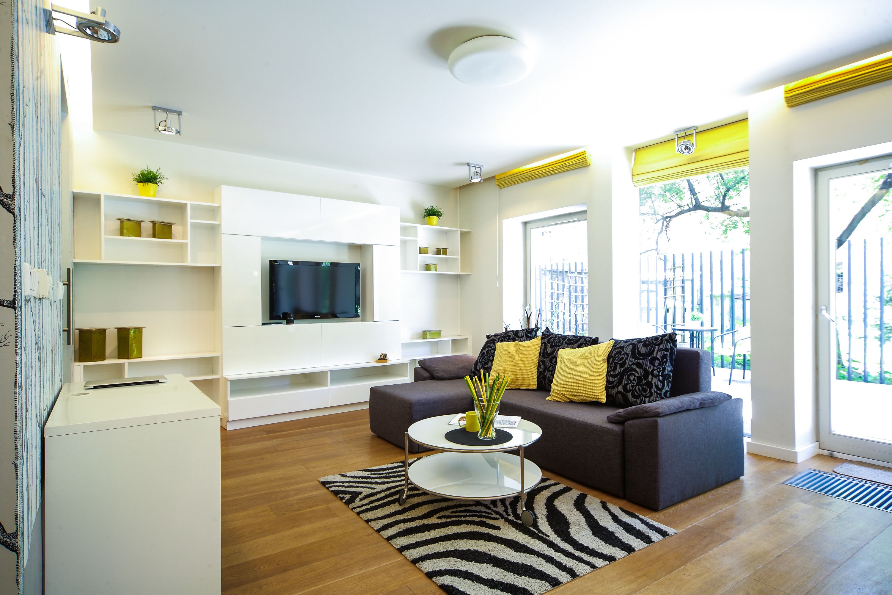 A modern living room with a sofa, coffee table, and TV. There are shelves with decorative items and a window with a view.