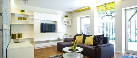 A modern living room with a sofa, coffee table, and TV. There are shelves with decorative items and a window with a view.