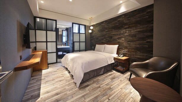 A modern bedroom with a wooden floor, a bed with white bedding, a wooden nightstand, a leather chair, and a wall-mounted TV. - Micro Hotel (Taichung)