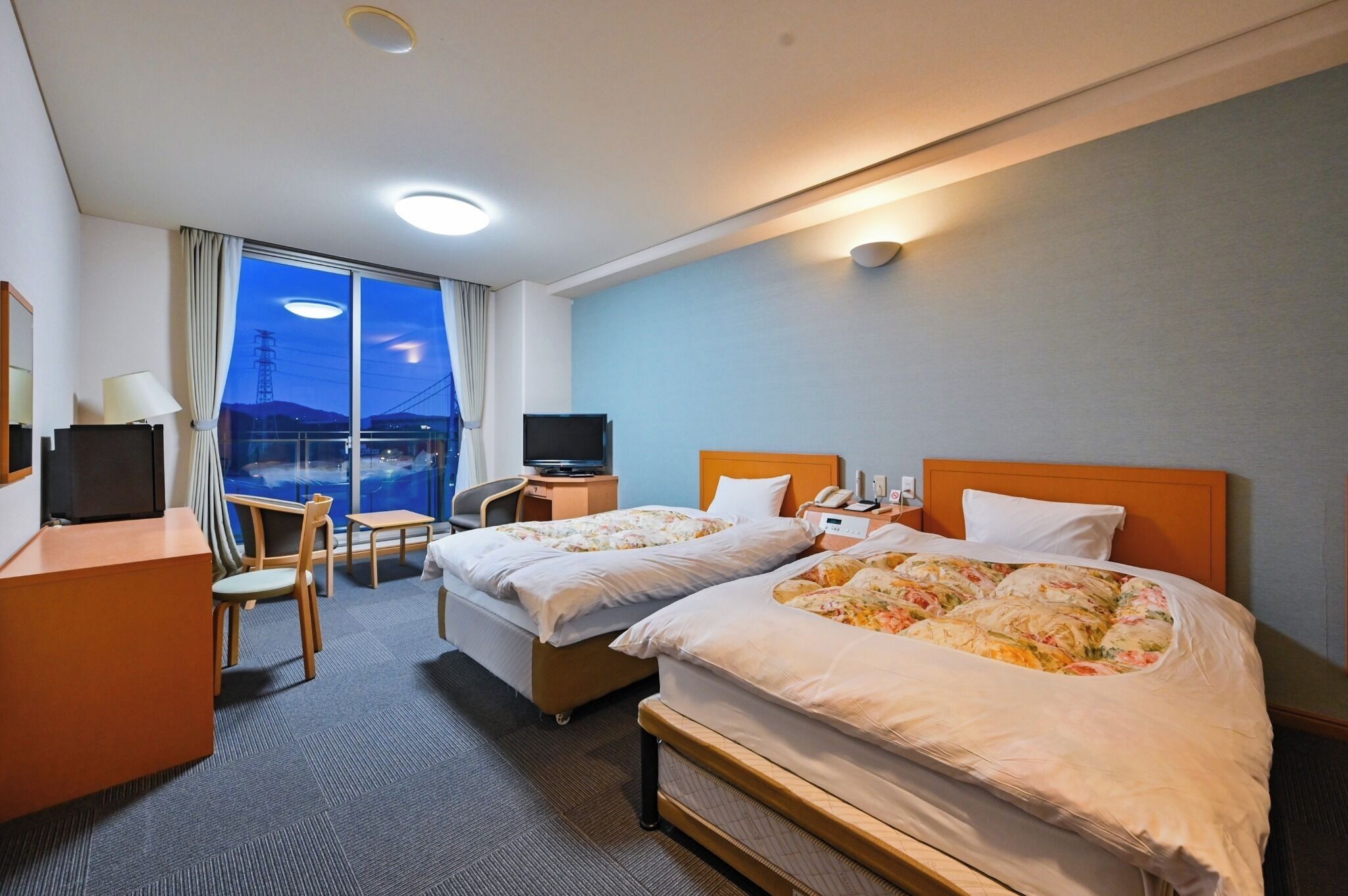 A hotel room with two beds, a desk, a chair, a TV, and a view of the outside.