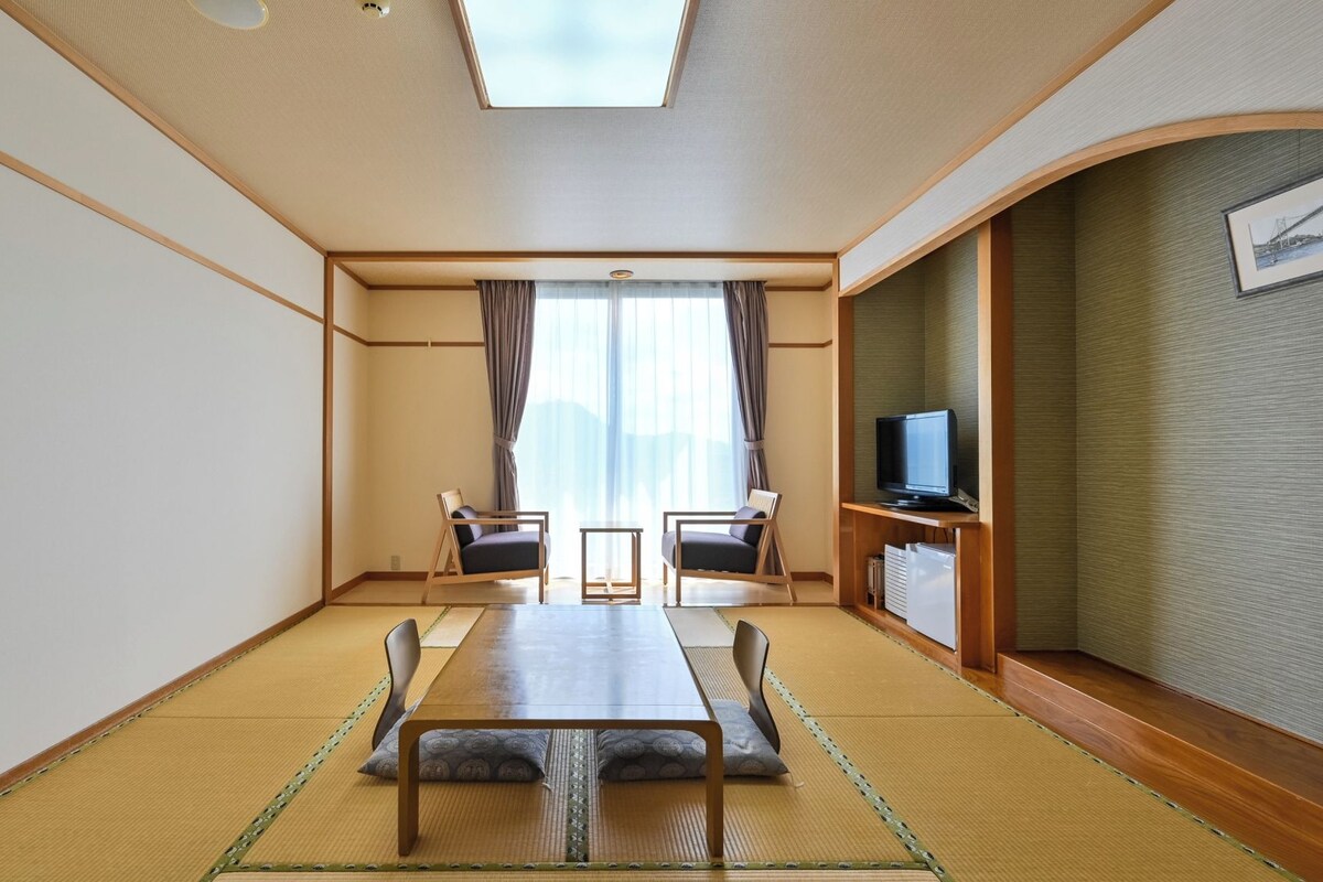 A traditional Japanese-style room with tatami flooring, a low table, two chairs, a television on a wooden stand, and a large window with curtains.