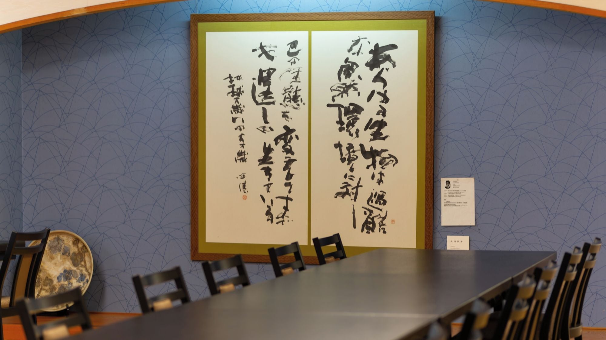 A conference room with a long table and chairs, a framed calligraphy piece on the wall, and a small informational card to the side.