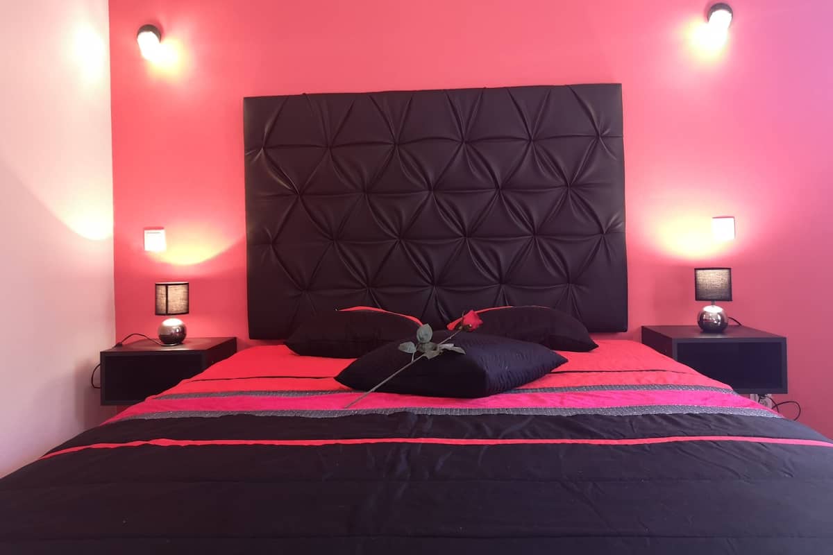 A bedroom with a tufted headboard, two bedside lamps, a black and red bedspread, and a single decorative pillow.
