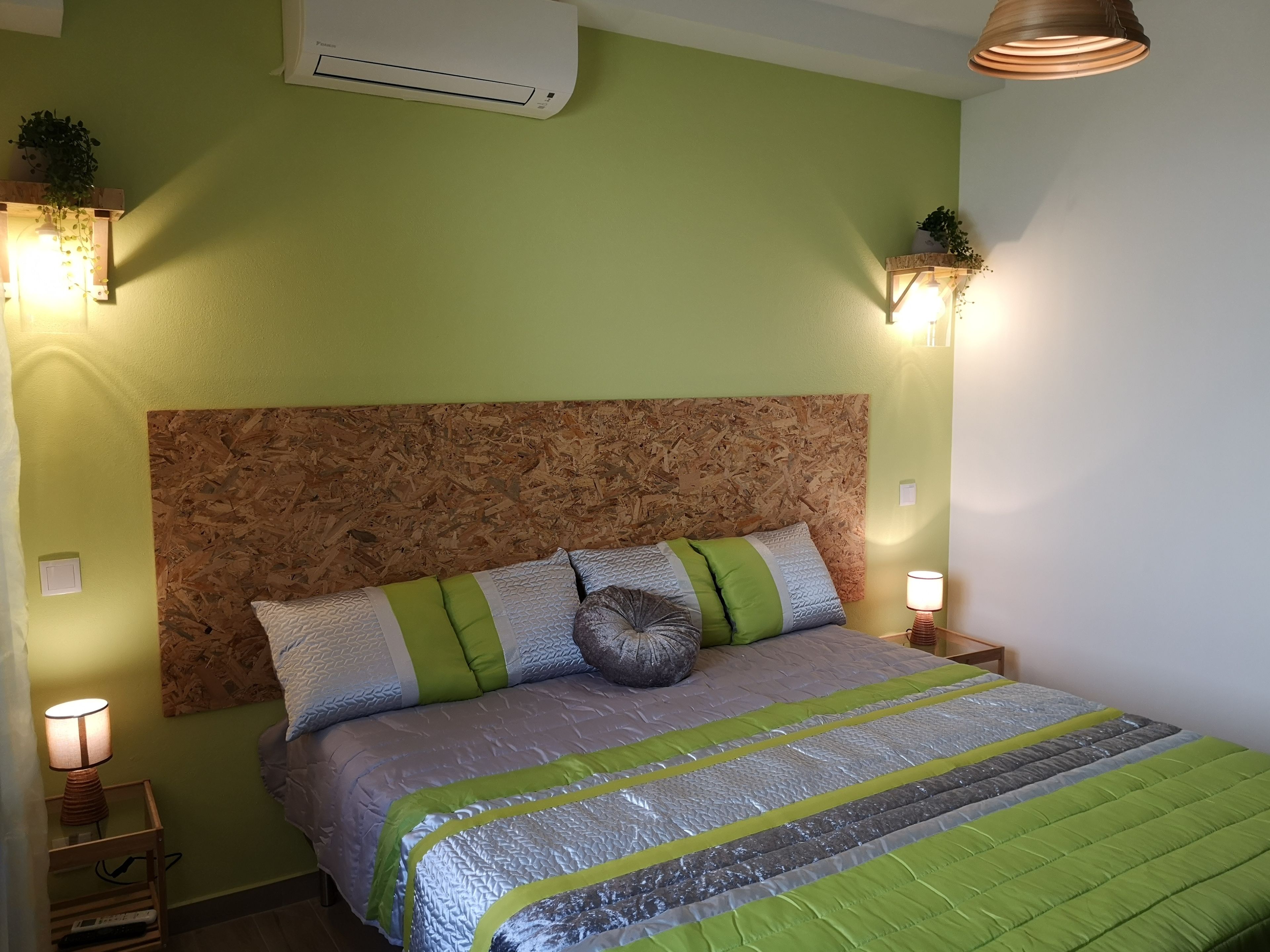 A bedroom with a wooden headboard, a bed with green and gray bedding, bedside lamps, and an air conditioner on the wall.