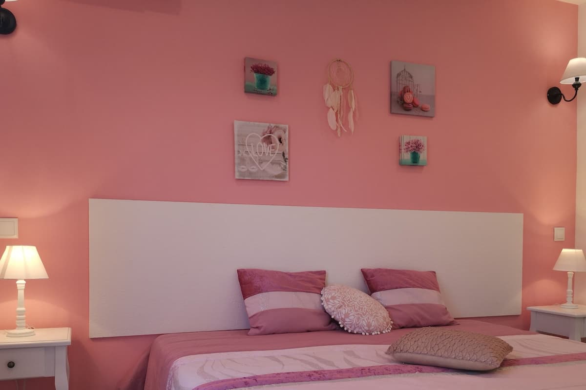 A bedroom with a pink wall, a white headboard, and two bedside tables with lamps. There are three framed pictures on the wall.